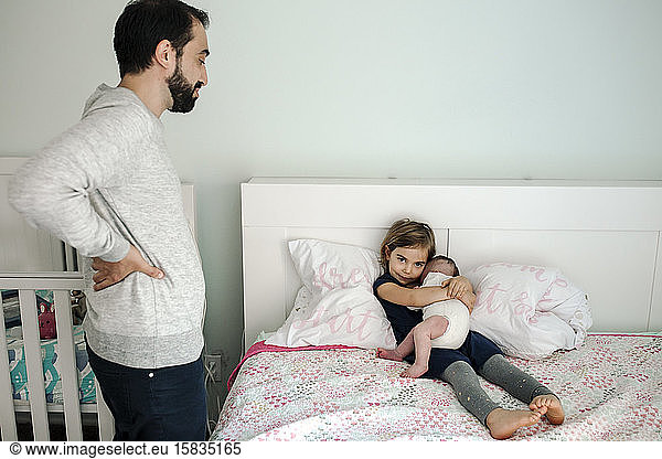 Bearded dad watching 4 yr old daughter on bed holding newborn