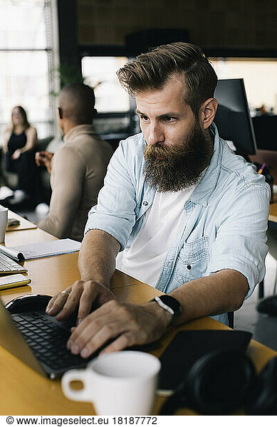 Bearded male computer programmer working on laptop at desk in office