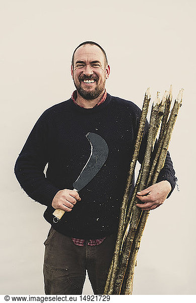 Bearded man holding bill hook and bunch of wooden stakes for traditional hedge laying  smiling at camera.