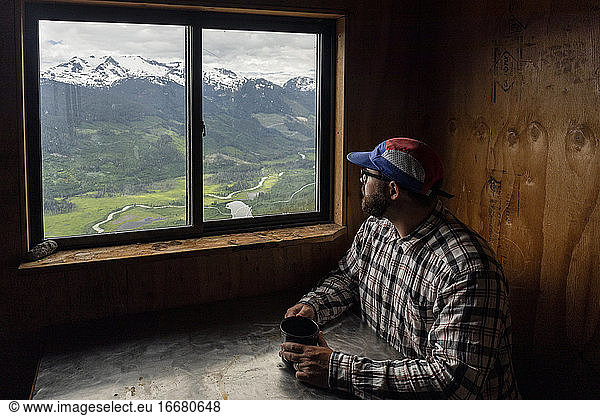 Bearded man in checkered shirt and cap sitting at table with mug of hot beverage and looking at mountains while resting in cozy cabin in British Columbia  Canada