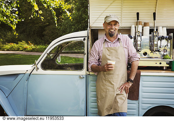 Bearded man wearing apron standing by blue mobile coffee shop  holding hot drink  smiling at camera.