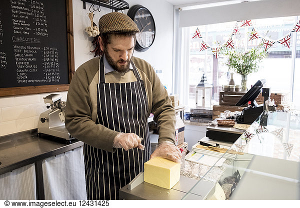 Bearded man wearing flat cap and apron standing at counter in a delicatessen  cutting cheese.