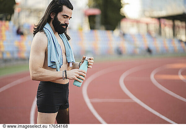 Bearded shirtless athlete holding bottle standing on running track