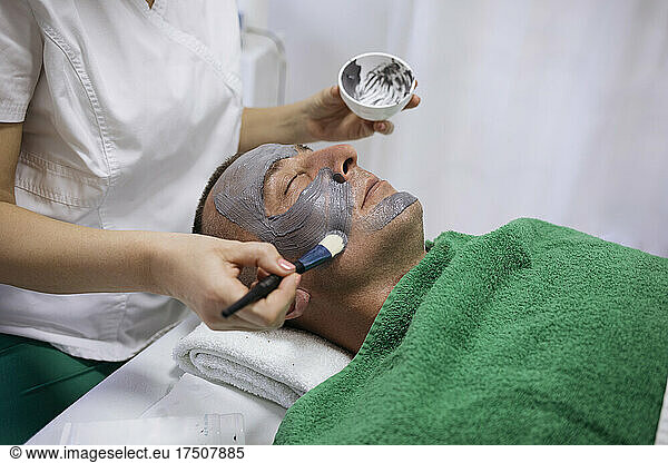 Beautician applying facial mask with brush on customer's face in beauty salon