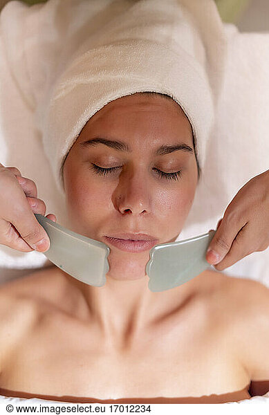 Beautician holding gua sha on female customer's chin at health spa