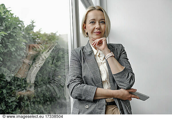 Beautiful blond businesswoman standing with hand on chin by glass window in office