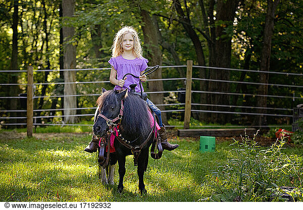 Beautiful blond girl in purple shirt riding a little black pony.