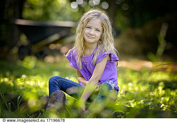 Beautiful little blond girl in purple shirt sitting on the grass.