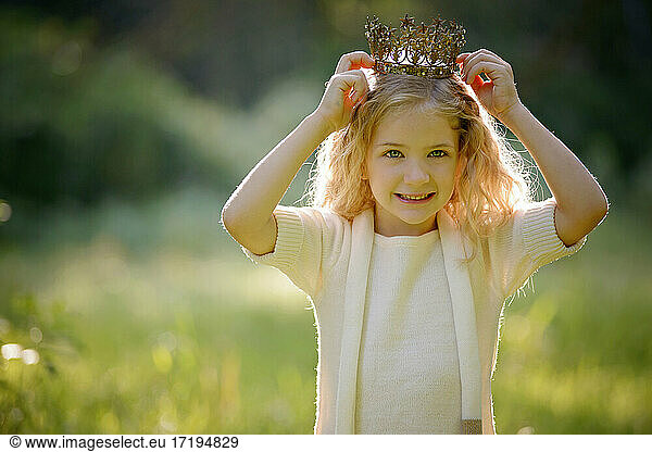 Beautiful little blond girl putting on a crown outdoors.