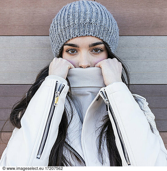 Beautiful teenage girl in knit hat covering her mouth with sweater against wall