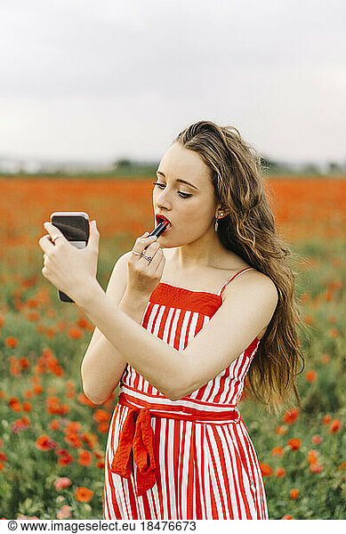 Beautiful woman applying lipstick standing at poppy field