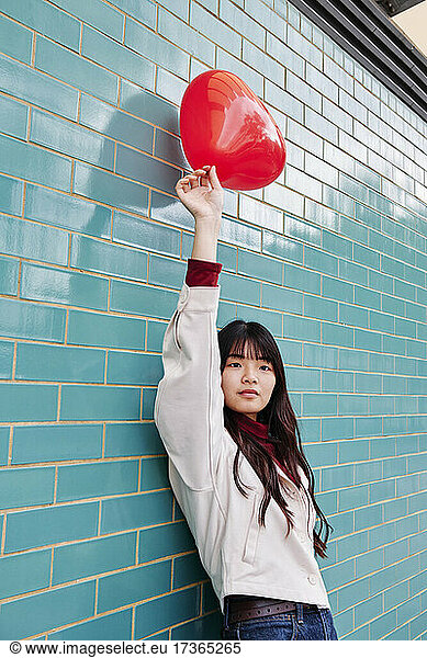 Beautiful woman holding red balloon while leaning on turquoise brick wall