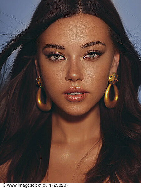 Beautiful woman smooth long hair brunette evening make up with liner tanned skin beautiful female portrait over blue background wearing gold earrings