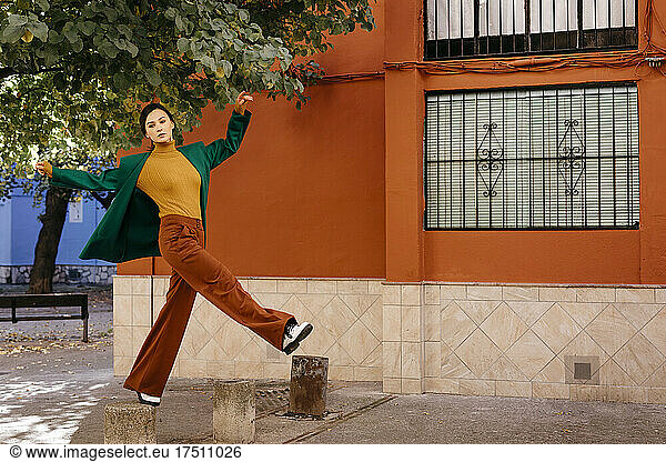 Beautiful woman wearing green jacket climbing on bollard in city