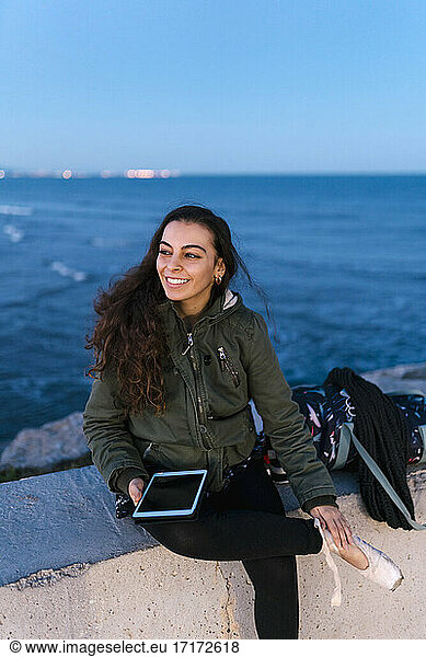 Beautiful woman with digital tablet sitting on retaining wall at beach during dusk against sky