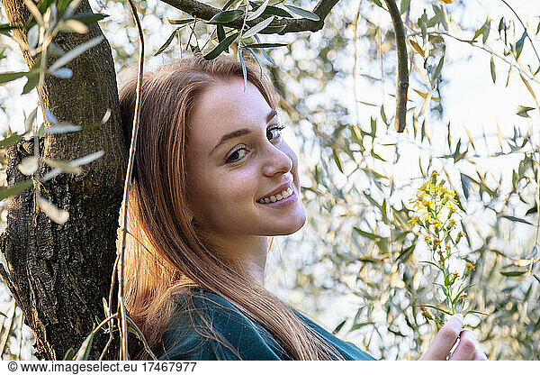 Beautiful woman with wildflower leaning on olive tree