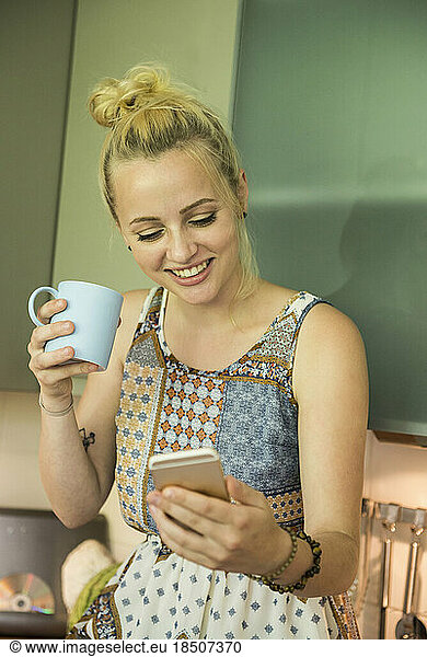 Beautiful young woman looking at smartphone and drinking coffee in the kitchen  Munich  Bavaria  Germany
