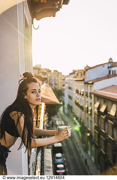 Beautiful young woman on balcony above the city at sunset with cup of coffee