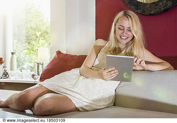 Beautiful young woman using digital tablet and sitting on sofa in the living room  Munich  Bavaria  Germany