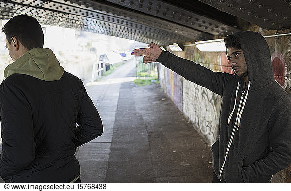 Bedrohlich wirkender junger Mann  der in einem städtischen Tunnel eine Fingerpistole auf einen Mann richtet