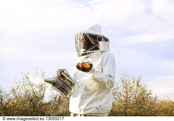 Beekeeper holding smoker and honey in jars while standing against sky