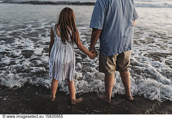 Behind view of red headed daughter holding father's hand in ocean
