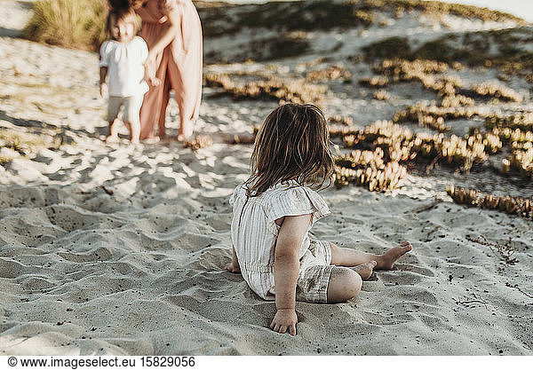 Behind view of young toddler girl sitting in sand looking at family