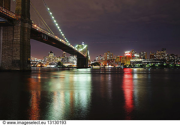 Beleuchtete Brooklyn Bridge über den Fluss gegen die beleuchtete Stadt