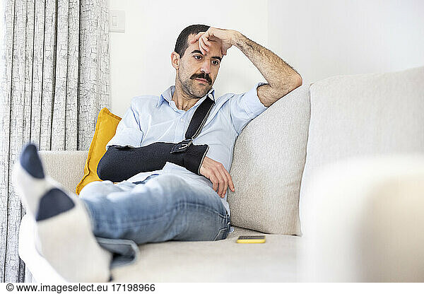 Besorgter Mann mit Armschlinge sitzt mit dem Kopf in der Hand auf dem Sofa zu Hause