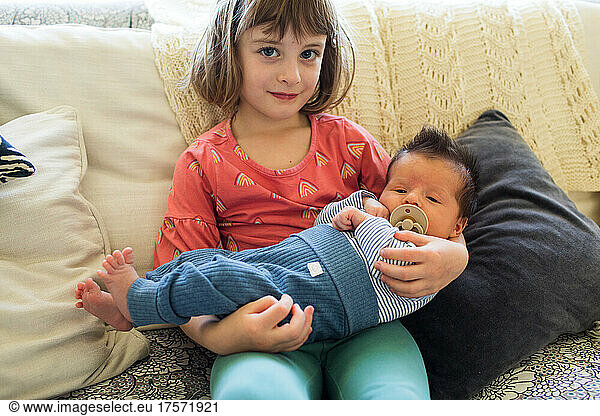 big sister holding hew newborn baby brother at home.