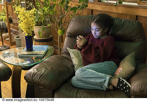 Biracial eleven year-old girl sitting on armchair staring at phone