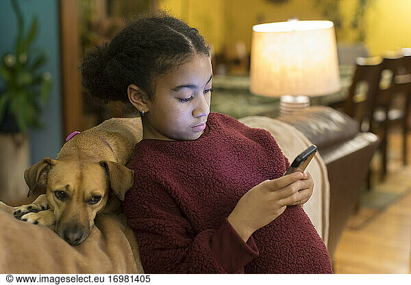 Biracial tween looking at phone while leaning against small brown dog