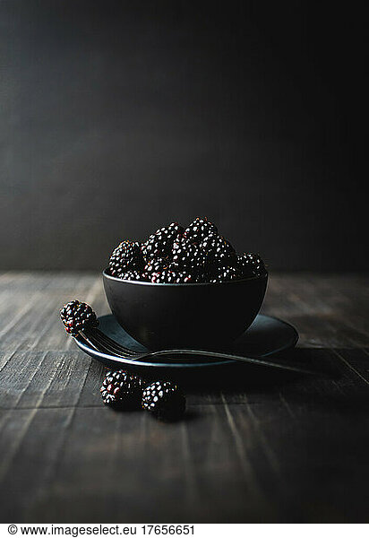 Black bowl of blackberries on wooden table with black background.