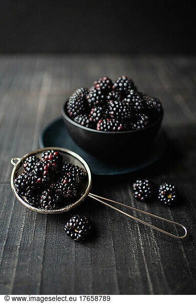 Black bowl of blackberries on wooden table with black background.