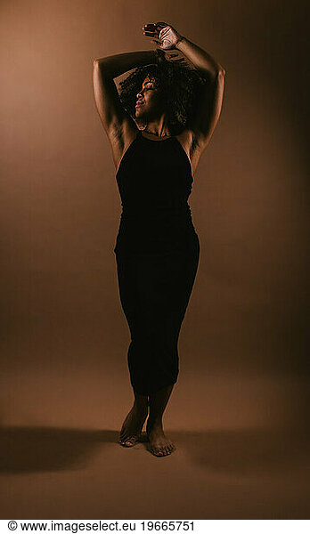 Black dancer pose arms over head on brown backdrop in figure dress