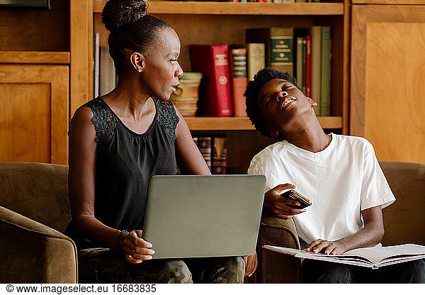 Black mom with computer homeschooling tired preteen son holding iPhone