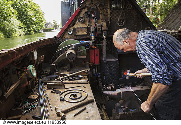 Blacksmith working in a small space on his narrowboat  a barge on river  bending over the anvil and shaping hot metal.