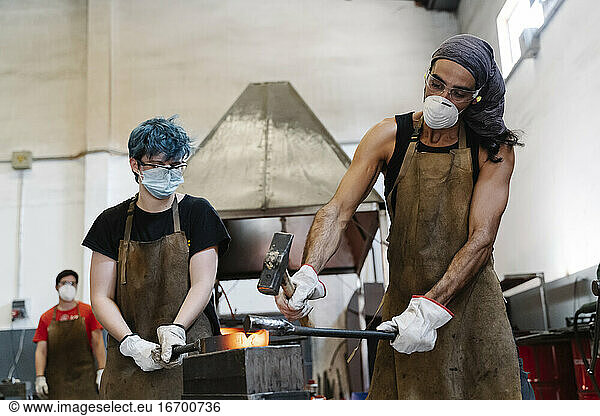 Blacksmiths in masks hitting hot metal with hammer while working