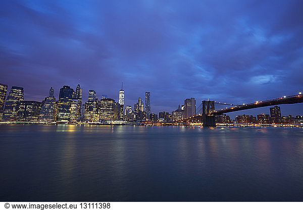 Blick auf die beleuchtete Skyline und die Brooklyn Bridge bei bewölktem Himmel in der Nacht
