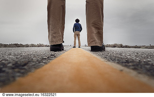 Blick durch stehende Beine auf einen lockigen Mann  der auf dem Mittelstreifen einer Straße steht
