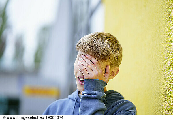 Blond boy covering face with hand in front of yellow wall