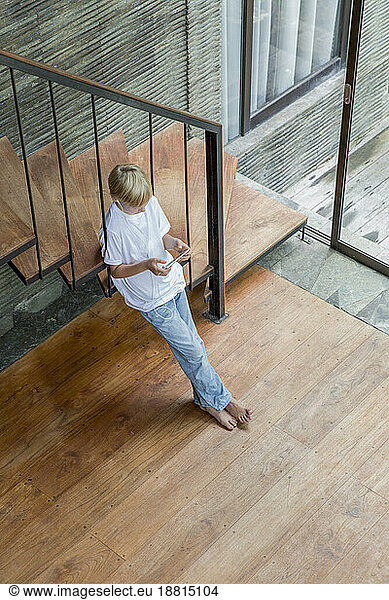 Blond boy leaning on staircase and using smart phone at home