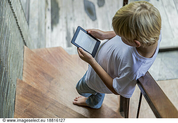 Blond boy using e-reader and standing on staircase at home