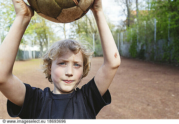 Blond boy with bruise on forearm holding soccer ball on playground