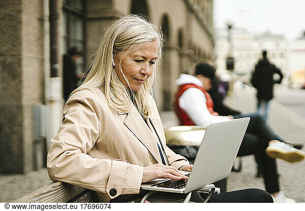 Blond businesswoman using laptop while sitting on bench in city