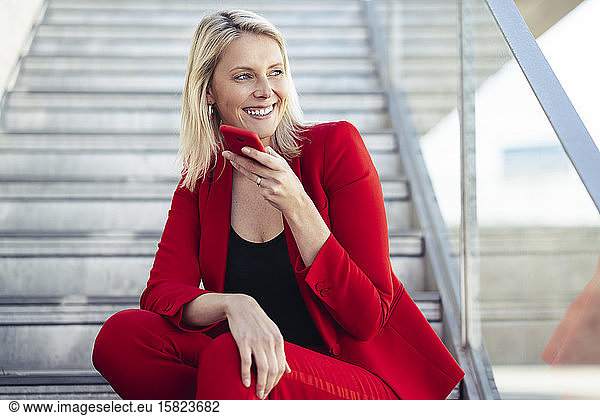 Blond businesswoman wearing red suit and using hands free telephone  sitting on stairs