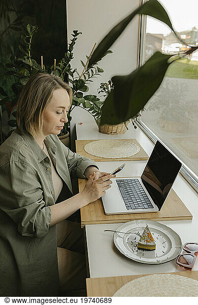 Blond freelancer using smart phone and sitting with laptop in coffee shop