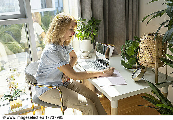 Blond freelancer writing in dairy sitting on chair at desk in home office