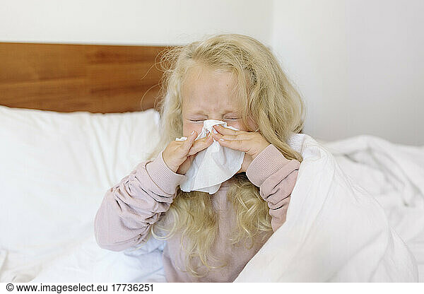Blond girl blowing nose with tissue paper in bedroom