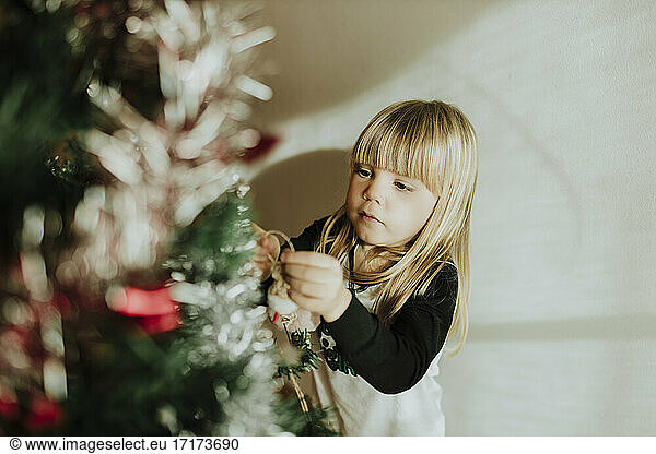 Blond girl decorating Christmas tree against wall at home